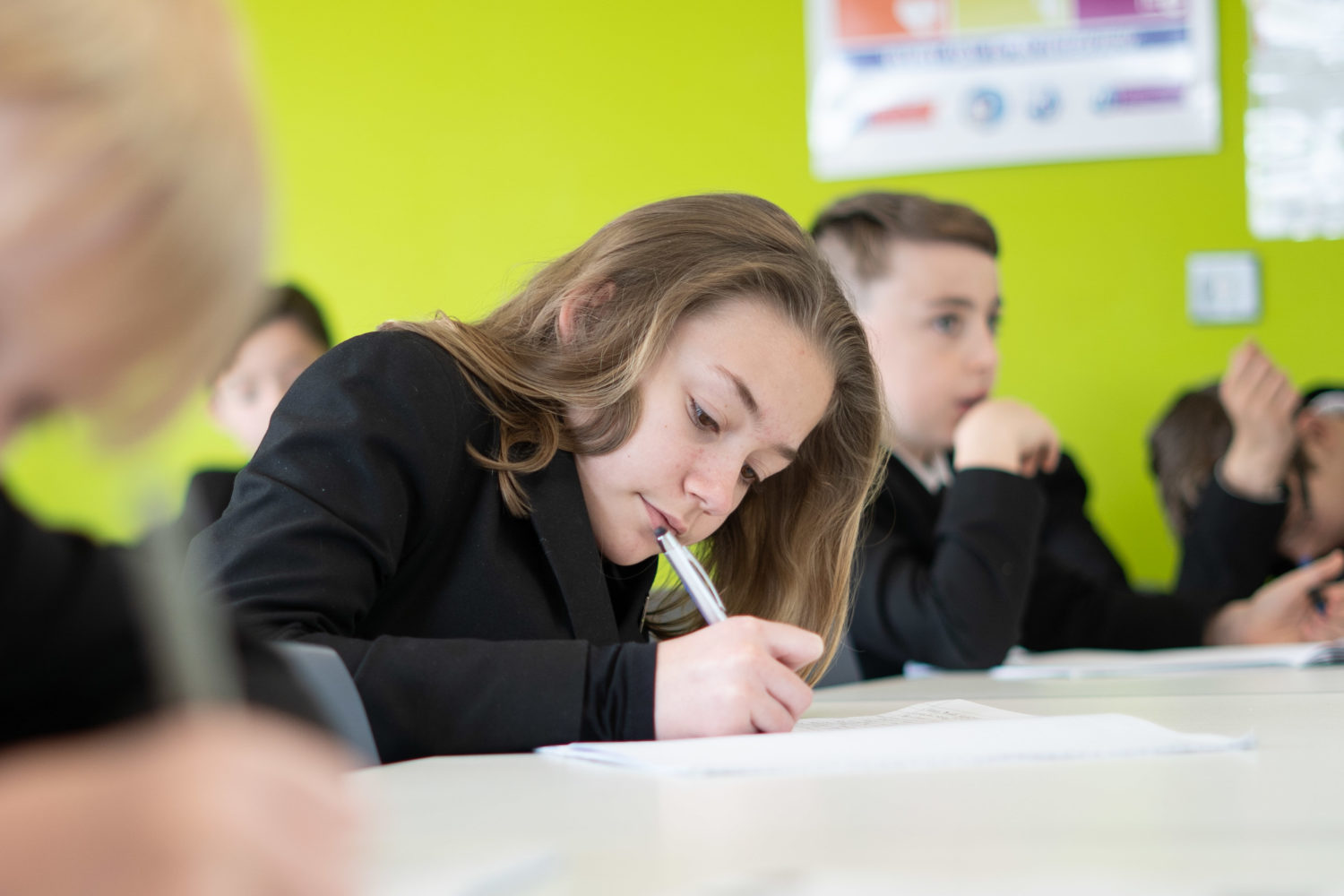 Some Leigh UTC students are shown sitting at a desk in a classroom, writing with pens in their hands.