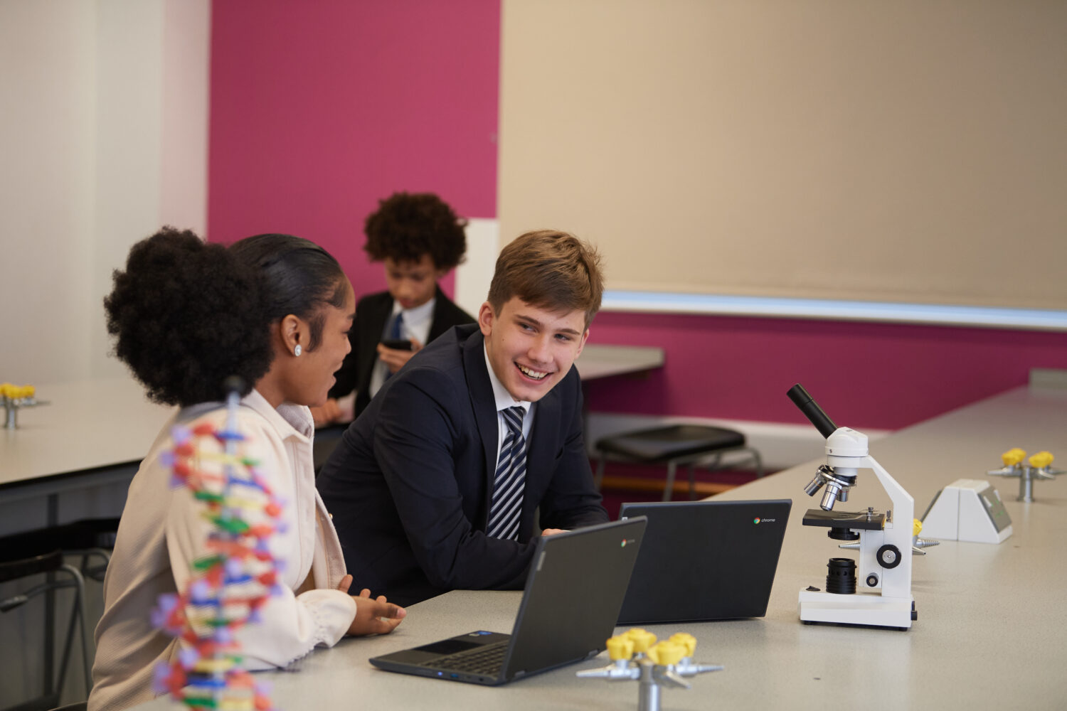 A male student is pictured sat at his desk, smiling. A female member of staff is seen sat beside him, assisting him with his learning.
