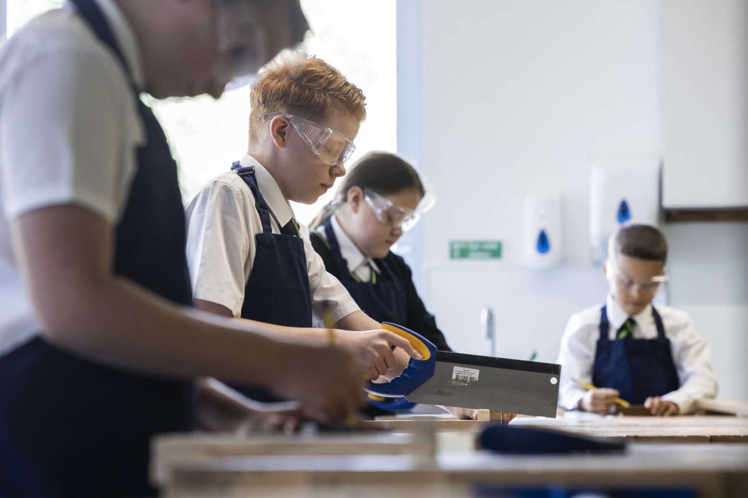 Four students, three boys and a girl, are pictured wearing safety goggles and using saws to cut materials in a DT class.
