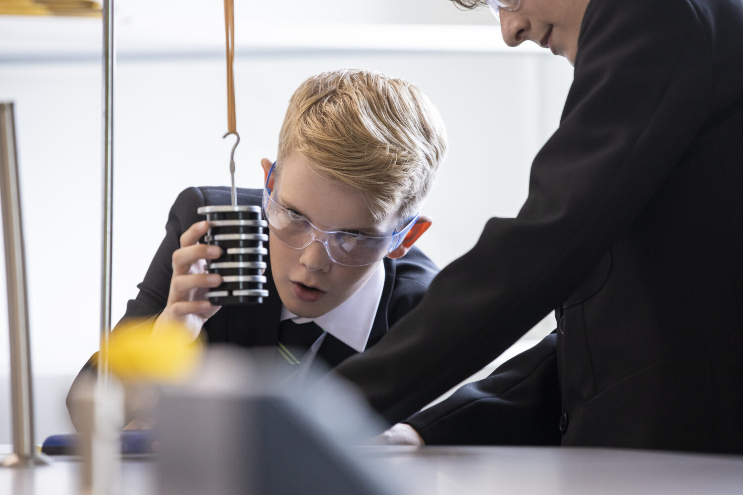 Two male students are pictured conducting a Science experiment together using weights and scales, whilst wearing safety goggles.
