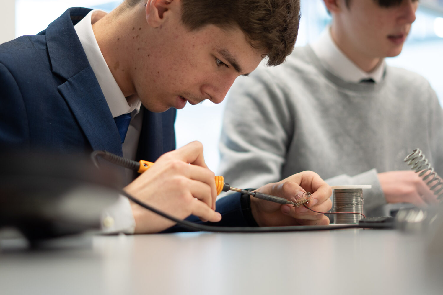 Two male Post-16 students can be seen sat at their desks, using soldering irons to combine metal objects together.