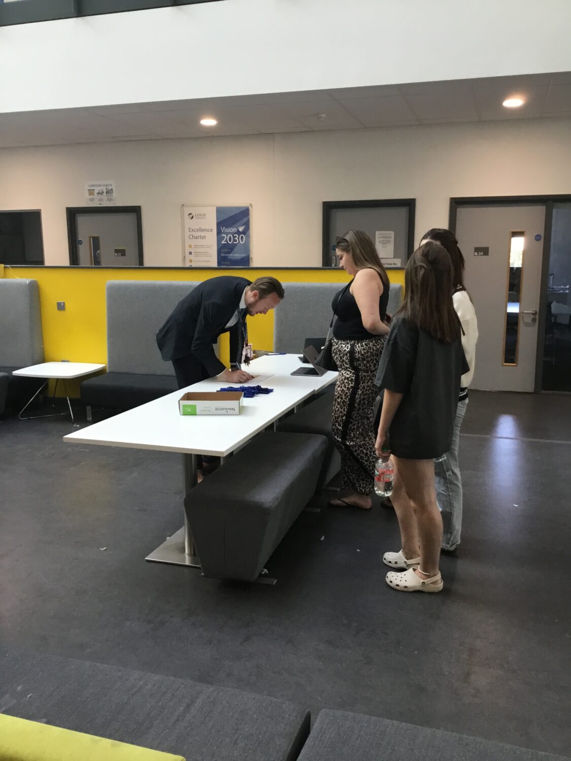Some Post-16 students are pictured collecting their A-Level results from a member of staff behind a desk.